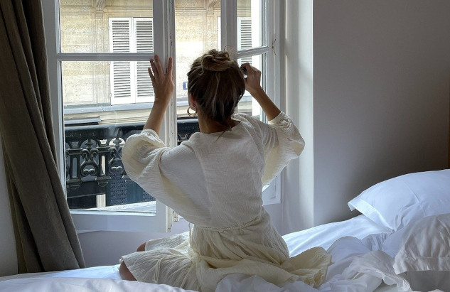 woman sitting on the edge of her bed facing an open window